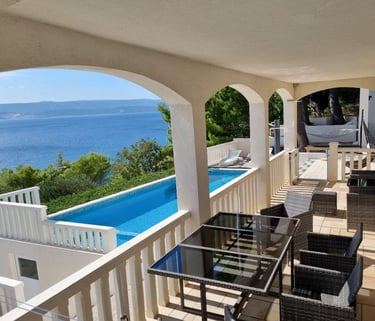 The photo shows an apartment balcony with a dining set and a view of the pool and the sea.