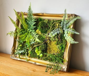 Preserved moss wall art with green ferns in an ornate gold vintage picture frame on a wooden table.