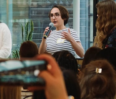 Photo of Micha in a stripy t-shirt holding a microphone and speaking to an audience