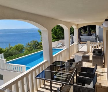 The photo shows an apartment balcony with a dining set and a view of the pool and the sea.