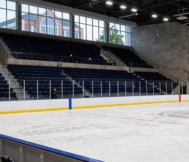 A crisp, wide-angle shot of a modern collegiate ice hockey rink in a North American / US Southern university campus setting. The lighting is bright and professional, highlighting the dark navy blue and royal blue accents of the stadium seating. In the background, students walk past large glass windows showing the architectural style of a Southern educational institution.