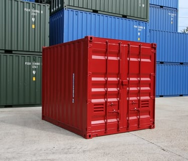 Small red steel shipping container in a shipyard with stacked blue and green cargo containers.