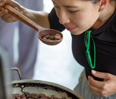 A woman smells roasted cacao beans in a wooden spoon over a large pan during traditional chocolate production.
