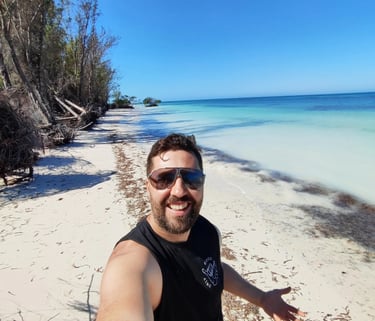 Smiling man taking a selfie on a tropical white sand beach with turquoise ocean water and clear blue sky.