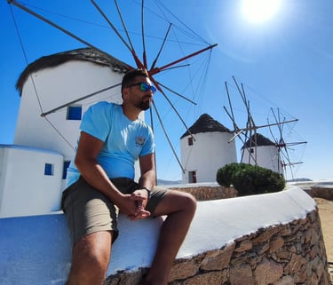 A man sitting on a stone wall in front of the historic white Mykonos windmills under a bright sun.