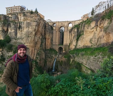 A smiling man stands before the historic Puente Nuevo bridge and waterfall in Ronda, Spain.