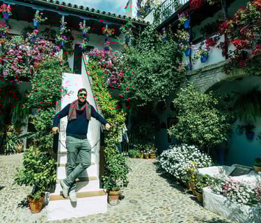 A man stands on stairs in a lush Cordoba patio filled with blooming flowers and hanging pots.
