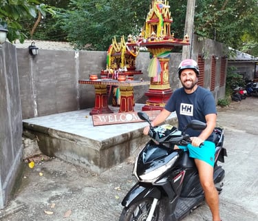 A smiling man wearing a helmet sits on a black scooter near a traditional Thai spirit house.
