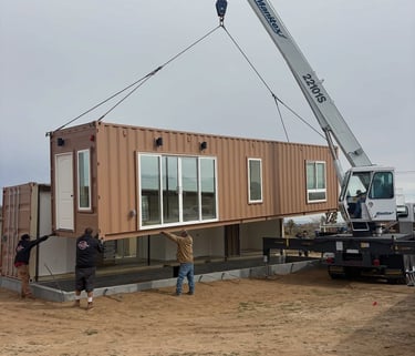 A construction crane lifts a modular shipping container home onto a foundation at a build site.