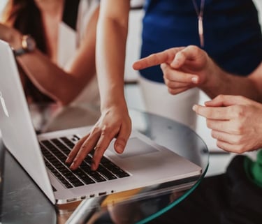 Personas colaborando en una mesa de cristal, señalando y escribiendo en un portátil.