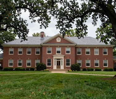 A wide photography shot of a North American / US traditional brick building used for education, surrounded by tall oak trees in a lush green lawn.