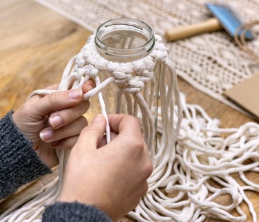 Manos tejiendo macramé con cordón de algodón sobre un frasco de vidrio.