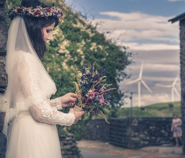 A bride in a lace wedding dress holding a wildflower bouquet and wearing a floral crown outdoors.