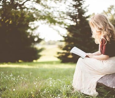 Person with long blonde hair reading a book on a sunlit rock in a grassy, wooded area.