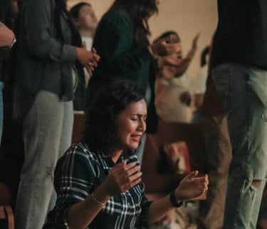 Person kneeling with eyes closed and hands raised, surrounded by others in communal worship.