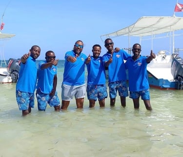 A group of smiling scuba intructors in blue shirts giving thumbs up in the ocean near boats.
