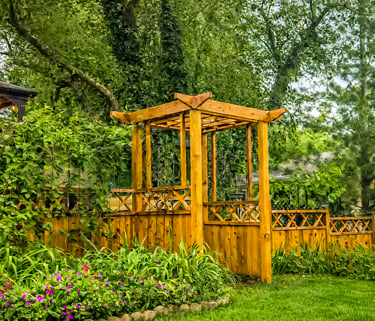 A wooden garden pergola and fence surround a lush backyard with green plants and purple flowers.