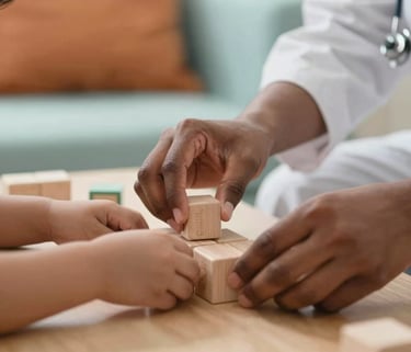 A close-up of a South Asian therapist's hands gently guiding a child's hands over a set of educational blocks, warm indoor lighting, soft focus background with muted orange and light teal accents.
