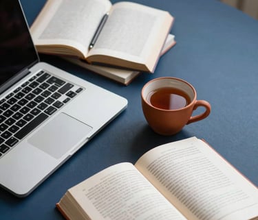 A high-angle shot of a writer's desk with a laptop, several open books, and a terracotta clay cup of tea, set against a dark blue background.
