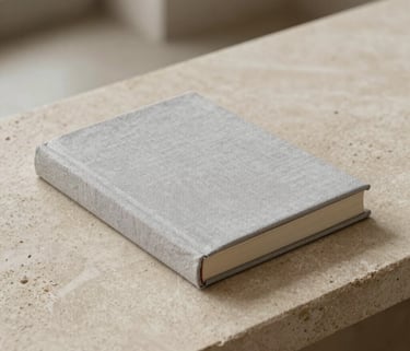 Minimalist photography of a single book bound in light grey linen resting on a sand-colored stone surface, soft natural light, South American / Spanish study interior, clean and artistic composition.