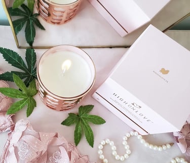 The photo shows a HighOnLove product  - a massage candle - on a background with some pearls, hemp leafs, and the packaging.