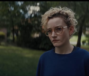 Julia Garner stands solemnly in a navy sweater and round glasses, framed by a suburban neighborhood