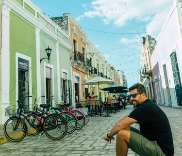 A man sits on a cobblestone street lined with colorful colonial buildings and parked bicycles in Campeche, Mexico.