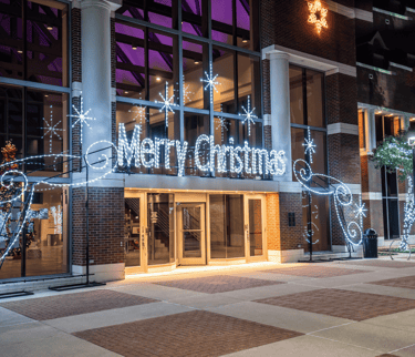 Merry Christmas neon sign and white holiday lights decorating a brick building entrance at night.