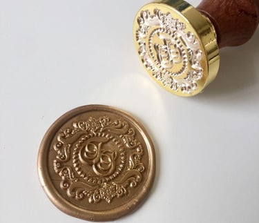 Custom brass wax seal stamp and a finished gold wax seal with an ornate monogram design on white paper.