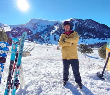 Smiling male skier in yellow jacket standing on a snowy mountain slope with blue skis under a bright sun.