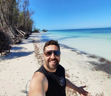 A smiling man takes a selfie on a sunny tropical white sand beach with turquoise ocean water.