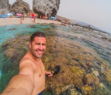 Smiling man metal detecting in clear ocean water at a rocky Mediterranean beach resort.