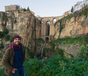 A smiling man stands before the historic Puente Nuevo bridge and waterfall in Ronda, Spain.