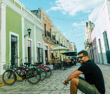 A man sits on a cobblestone street lined with colorful colonial buildings and parked bicycles in Campeche, Mexico.