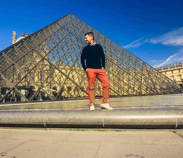 A man posing in front of the Louvre Pyramid in Paris, France, on a sunny day.