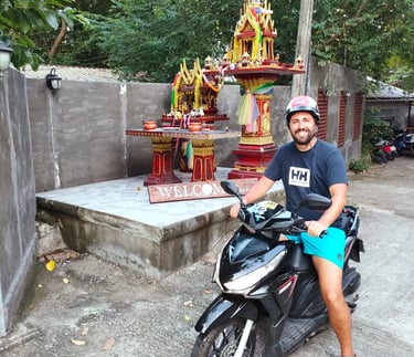 A smiling man wearing a helmet sits on a black scooter in front of a colorful Thai spirit house.