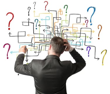 A man in a suit faces a whiteboard covered with a complex web of arrows, boxes, and colored question marks, seemingly perplex