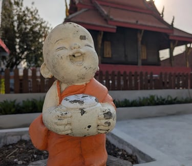 A smiling monk statue in orange robes at a Buddhist temple in Thailand during sunset.