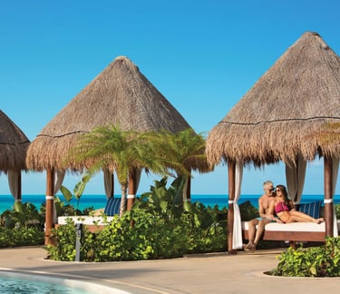 A couple relaxes in a luxury beachfront cabana with thatched roofs at a tropical resort.