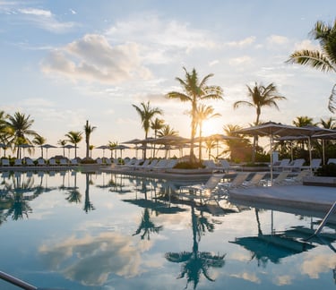 Luxury tropical resort pool with lounge chairs and palm trees reflecting in the water at sunset.