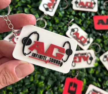 Custom 3D printed AG Infiniti Sound logo keychain held against a green hedge background.
