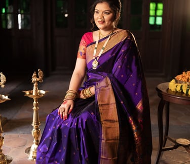 Indian woman in a purple Paithani silk saree with gold jewelry sitting in a traditional setting.