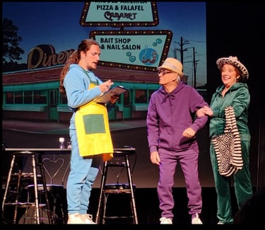 Actors in colorful tracksuits perform a comedic theater scene in front of a retro diner backdrop.