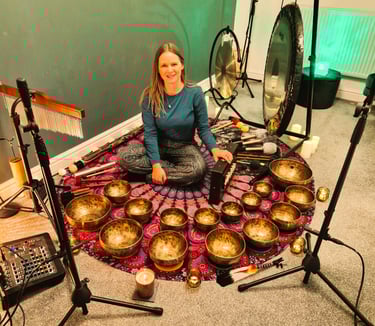 Chrissy sitting on a mat with shruti box, chimes, gongs, many singing bowls, and audio equipment