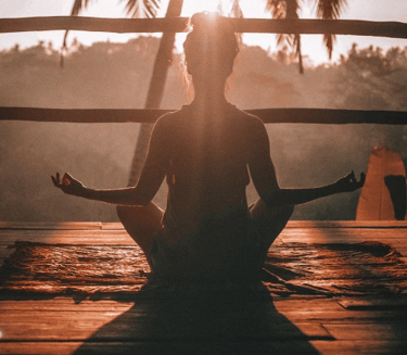 Silhouette of a woman practicing morning yoga meditation on a wooden deck during a tropical sunrise.