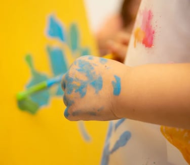 Child's hand covered in blue paint while creating artwork on a yellow canvas