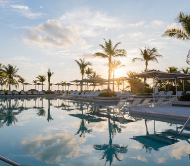 Luxury tropical resort pool with lounge chairs and palm trees reflecting in the water at sunset.