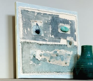 textured mixed media painting on canvas in muted denim blue tones, with layered stitched fabric and natural stone elements.