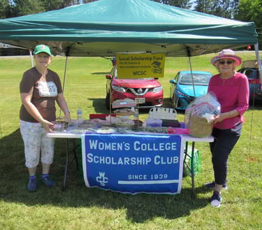 photograph of two women standing at a bake sale table for College Scholarship Club under small tent