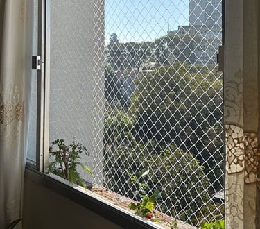 janela de apartamento vista por dentro com rede de proteção; cortinas claras nas laterais, plantas no parapeito e, ao fundo,